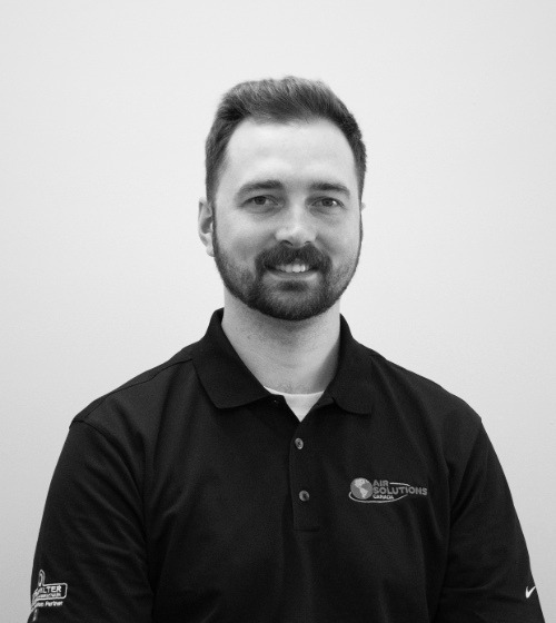 Man with short hair and a beard, wearing a black polo shirt with "Air Solutions" logo, sits against a plain light background, looking at the camera and smiling slightly.