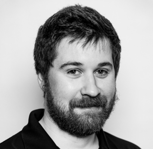 A bearded person with short hair, wearing a dark shirt, poses for a black and white portrait against a plain background.