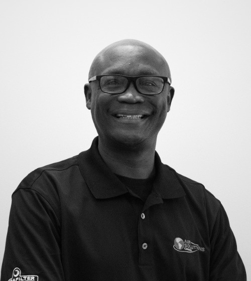 A man wearing glasses and a black polo shirt with an "Air Solutions" logo smiles while standing against a plain, light-colored background.