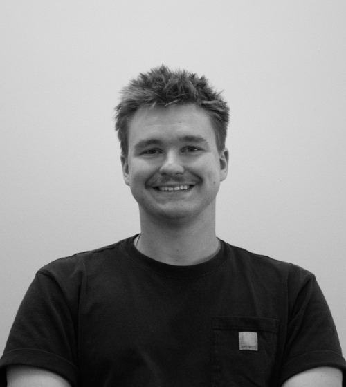 A young man with short, messy hair and a mustache is smiling, wearing a dark T-shirt, and standing against a plain, light-colored background.