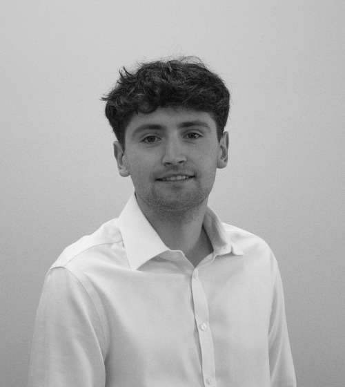 A young man with short, dark curly hair wearing a white button-down shirt stands against a plain light background, looking at the camera and smiling slightly.