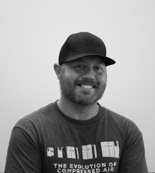 Bearded man wearing a dark baseball cap and graphic T-shirt smiles at the camera against a plain light background.