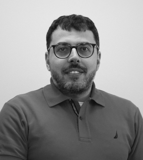 A man with short dark hair, glasses, and a beard is wearing a collared shirt and facing the camera against a plain light background.