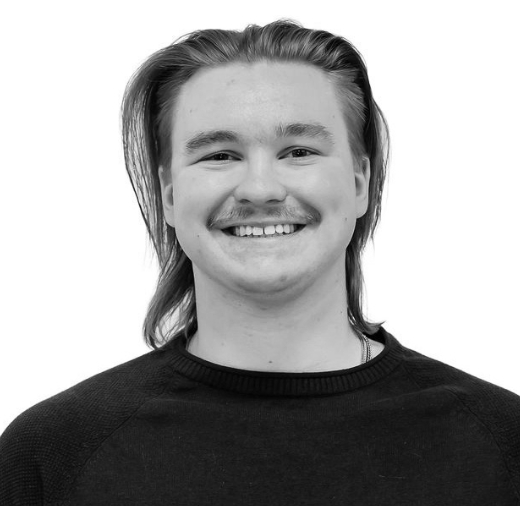 A person with long hair and a mustache, wearing a dark sweater, smiles against a plain white background.