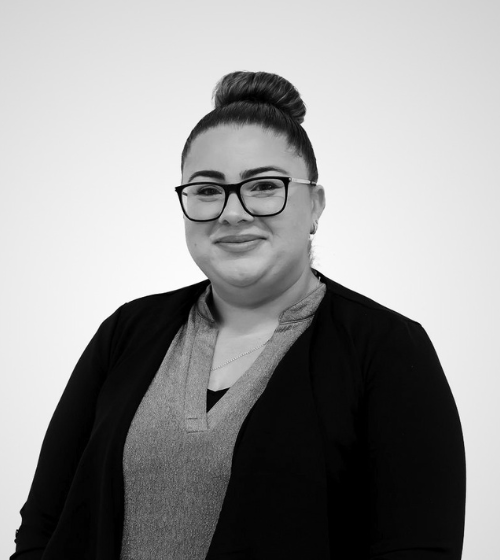 A woman with glasses and hair in a bun, wearing a light blouse and dark jacket, poses against a plain white background.