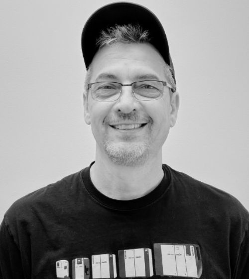 Man wearing glasses, a dark t-shirt with graphics, and a baseball cap stands in front of a plain light background, smiling at the camera. Black and white photo.