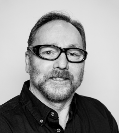 A man with a beard and glasses wearing a dark button-up shirt poses for a black and white portrait against a plain background.