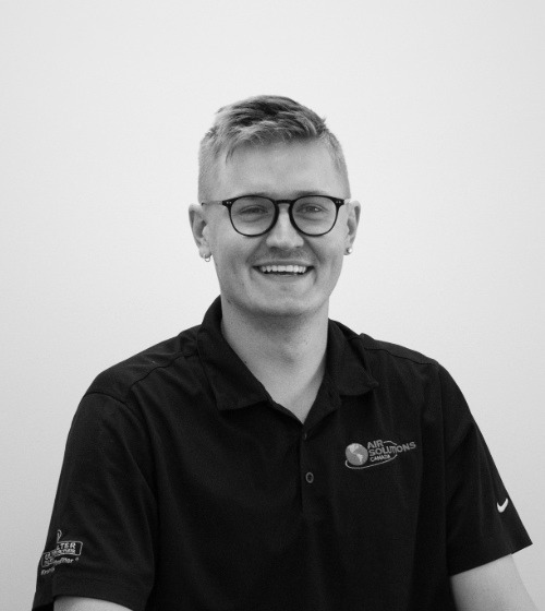 A person with short hair and glasses, wearing a black polo shirt with logos, sits smiling against a plain white background.