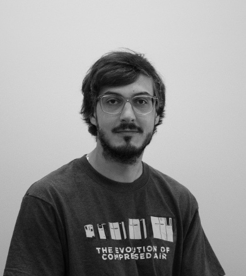 A person with glasses and a beard is sitting and facing the camera, wearing a T-shirt that says "THE EVOLUTION OF COMPRESSED AIR." The background is plain and light-colored.