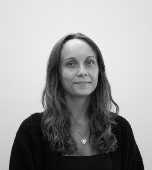 A woman with long wavy hair and a necklace, wearing a dark sweater, is standing against a plain light background and looking at the camera.